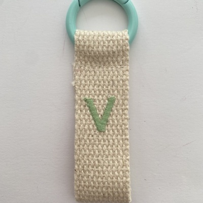 Pendurador de tecido bege com aro azul claro e letra V verde