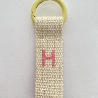 Porta-chaves com argola amarela e tira de tecido bege com letra H rosa bordada