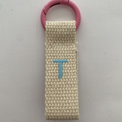 Porta-chaves com argola rosa e tira de tecido bege com letra T azul bordada