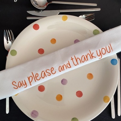 Prato branco com pontos coloridos, talheres de metal, guardanapo branco com bordado laranja e toalha preta com texto em português.