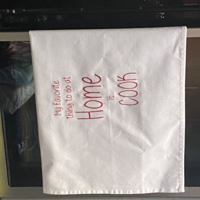 Pano de cozinha branco com texto bordado a vermelho pendurado em forno inox.