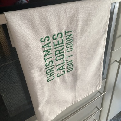 Pano de cozinha branco com texto verde bordado pendurado na porta de um forno.