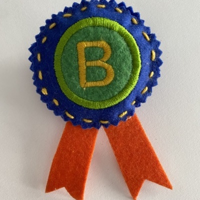 Insígnia de feltro azul com letra B verde e fita laranja