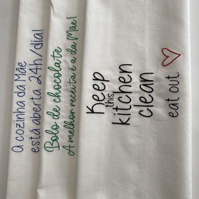 Três toalhas de cozinha brancas com bordados de textos em azul, verde e preto com coração vermelho.