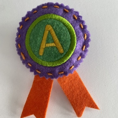 Distintivo de feltro roxo e verde com letra A e fitas laranja