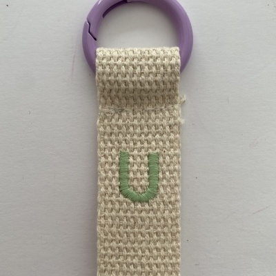 chaveiro com argola roxa e tira de tecido bege com letra U bordada a verde