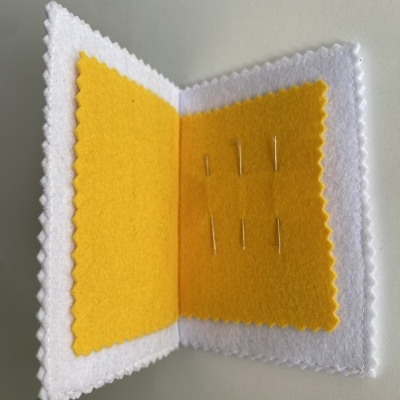 Estojo feltro branco aberto com interior amarelo e alfinetes prateados.