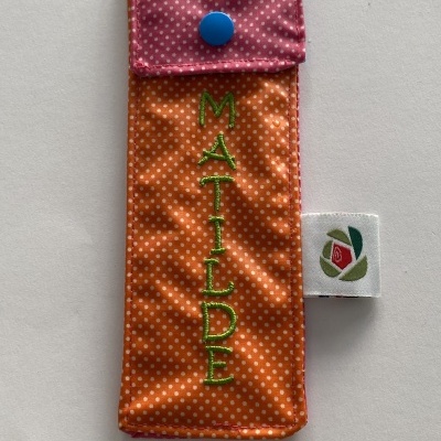 Estojo de tecido laranja com pontos brancos, tampa rosa com pontos e botão azul, nome Matilde bordado a verde.