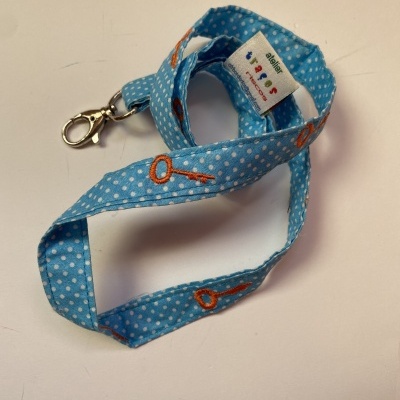 Cordão azul claro com bolinhas brancas e desenhos laranja, presilha metálica e etiqueta com texto