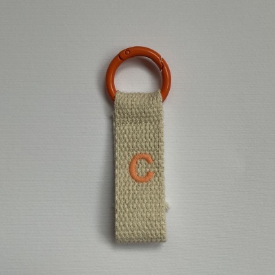 Porta-chaves com argola laranja e tira de tecido bege com letra C bordada