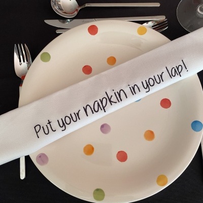Conjunto de louça com prato de pintas coloridas, guardanapo branco com texto em inglês, talheres e toalha preta bordada