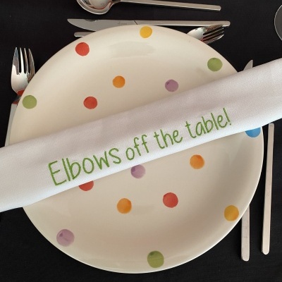 Conjunto de mesa com prato branco e pontos coloridos, guardanapo bordado, talheres aço inox e toalha preta com texto