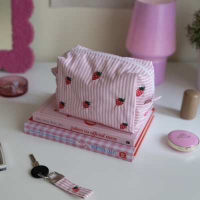 Necessaire rosa com padrão de morangos e riscas em cima de livros numa mesa branca com vários objetos