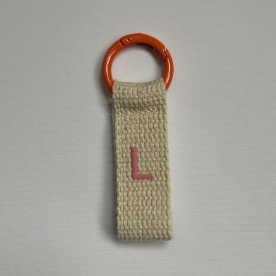 Porta-chaves com anel laranja e fita de tecido bege com letra L cor-de-rosa.