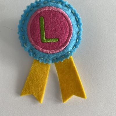 Distintivo de feltro azul e rosa com letra 'L' verde e fitas amarelas