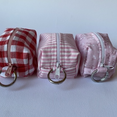 Três bolsas retangulares de tecido com fecho éclair e argola metálica prateada colocadas em linha sobre fundo branco.