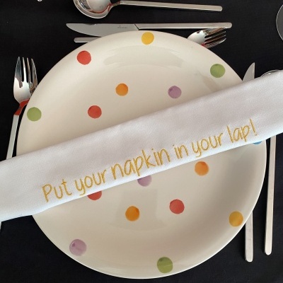 Mesa posta com prato bolinhas coloridas, talheres inox, guardanapo branco com texto em inglês e pano de mesa preto com texto em português