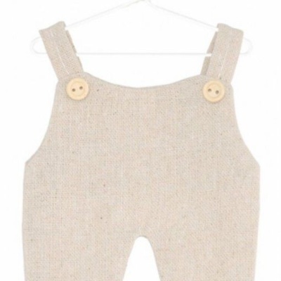 Macacão bege claro de bebé com alças e botões de madeira, pendurado num cabide branco