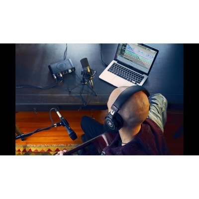 Mesa com laptop, interface de áudio, microfones, pessoa com auscultadores a tocar guitarra