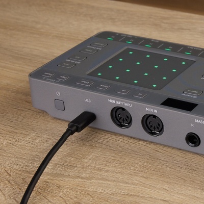 Controlador musical cinzento com pontos luminosos verdes e portas USB e MIDI numa mesa de madeira clara