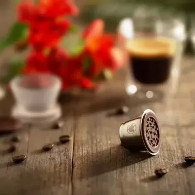 Cápsula metálica de café em mesa de madeira com café, grãos e flores vermelhas ao fundo