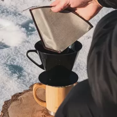 Filtro de café de tecido bege com bordas castanhas sobre suporte preto colocado numa caneca de madeira na neve.