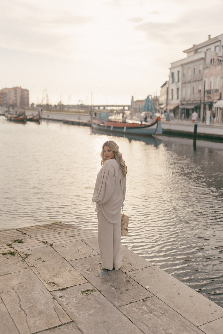 Blusa Serena Mulher loira com conjunto bege e bolsa de palha na margem de um canal com barcos e edifícios ao fundo