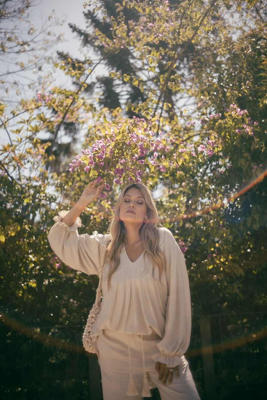Blusa Serena Mulher loira com roupa bege em ambiente exterior com árvores e flores lilases