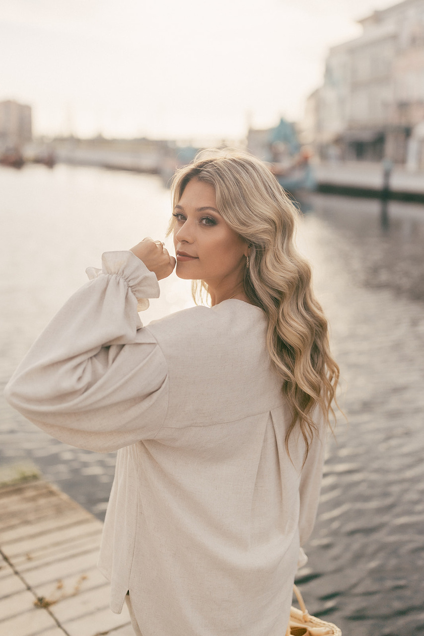 Blusa Serena Mulher com blusa creme de manga comprida junto a um cais com água e edifícios ao fundo