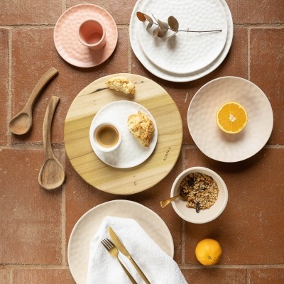 Conjunto de louças e talheres cerâmicos e de madeira para mesa, em tons claros, dispostos sobre chão de tijoleira