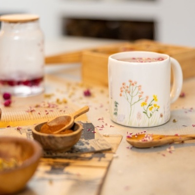 Caneca branca com padrão floral colorido e bebida com pétalas, colher e recipientes de madeira sobre mesa.