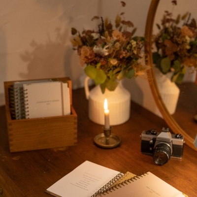 Mesa de madeira com caderno aberto, vela acesa, flores secas em vaso e câmara fotográfica em frente a espelho redondo
