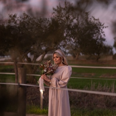 Mulher de vestido bege segurando bouquet de flores em cenário campestre ao pôr do sol
