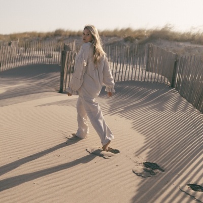 Mulher loira com roupa bege clara andando na areia junto a cerca de madeira ao pôr do sol