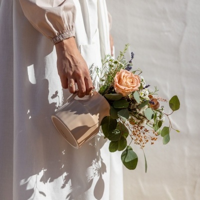 Pessoa com vestido branco segurando jarro bege com flores e folhagem