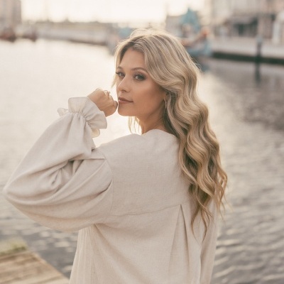 Mulher com blusa creme de manga comprida junto a um cais com água e edifícios ao fundo