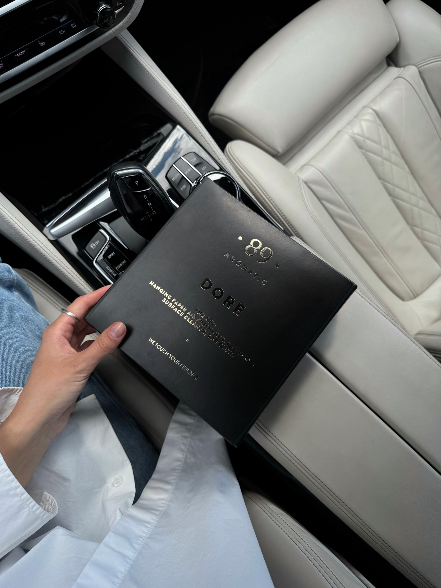 caixa preta com texto dourado na mão de uma pessoa dentro de um carro com bancos de couro branco