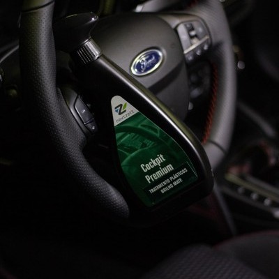 Produto de limpeza para automóveis 'Cockpit Premium' numa embalagem preta com rótulo verde, perto do volante de um carro Ford