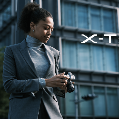 Mulher com fato cinzento segura câmara fotográfica diante de edifício moderno com texto X-T5