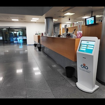 Quiosque interactivo branco com ecrã táctil azul e branco junto a balcão de madeira