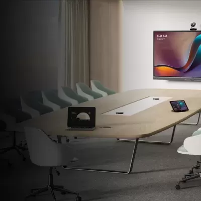 Sala de reunião com mesa oval, cadeiras brancas, monitor grande e dispositivos eletrónicos sobre a mesa