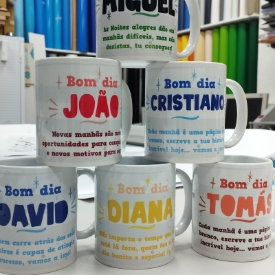 Canecas brancas com nomes e mensagens em várias cores empilhadas num espaço com materiais de escritório.