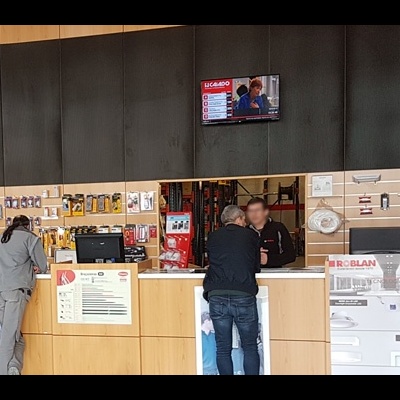 Mostrador de loja com acessórios pendurados, duas pessoas e ecrã que mostra canal 'LE CANARD'