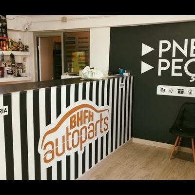 Interior de loja de peças automóveis com balcão às riscas, parede preta com texto PNEUS PEÇAS, cadeira preta e chão madeira