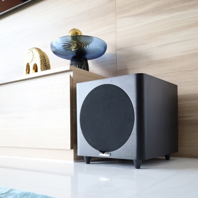 Subwoofer preto Cleveland com grelha frontal em tecido, em chão branco junto a mobília clara