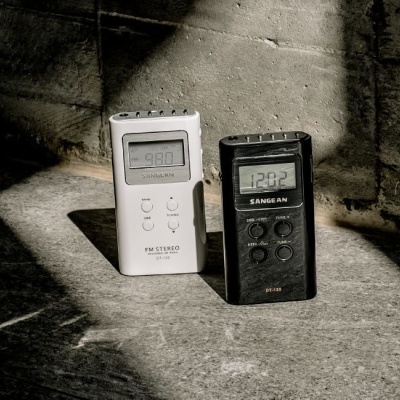 Dois rádios portáteis Sangean, um branco e outro preto, em chão de cimento