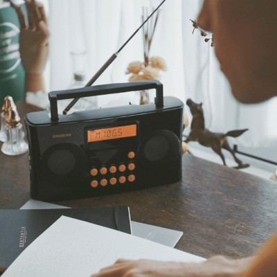 Rádio portátil preto com visor digital laranja e botões brancos numa mesa de madeira com objetos decorativos