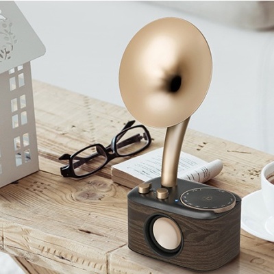 Coluna portátil retro em madeira com altifalante circular e tubo dourado ao lado de chávena, óculos e casa decorativa numa mesa