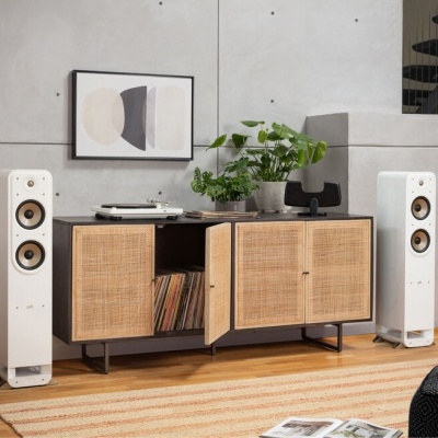 Sistema de som com colunas brancas, móvel com portas em palhinha, planta, quadro abstrato e tapete listrado