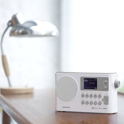 Rádio digital branco Sangean com ecrã e botões em cima de mesa de madeira com lâmpada ao fundo